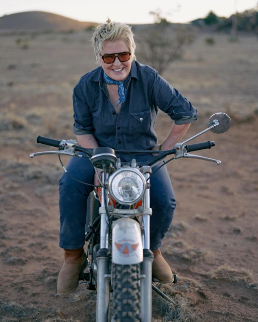 Adult woman with short blond hair is riding a vintage motorcycle in a rural desert setting with a warm color cast and diffused sunlight.