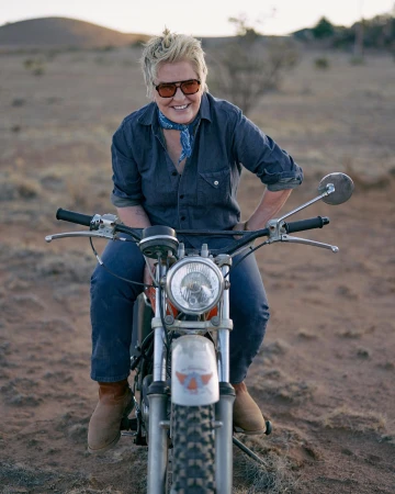 Adult woman with short blond hair is riding a vintage motorcycle in a rural desert setting with a warm color cast and diffused sunlight.