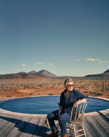 Person with short blonde hair seated on a wooden chair on a wooden deck next to an outdoor circular pool in a desert landscape