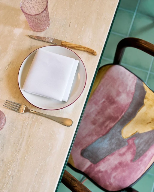 Overhead shot of a plate with white folded napkin, fork, knife, and pink patterned glass on a marble table with patterned chair.