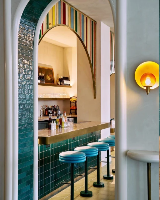 Interior view of an upscale bar with turquoise stools, a gold countertop, and an arched entryway with tiled accents and a golden light fixture.