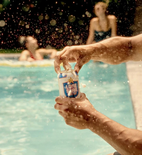 Close-up shot of hands opening a Lone Star beer can with water droplets and two females blurred in the background in a pool.
