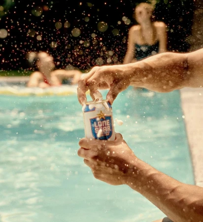 Close-up shot of hands opening a Lone Star beer can with water droplets and two females blurred in the background in a pool.