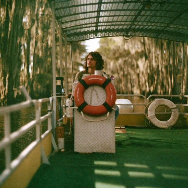 Adult male steers a pontoon boat in a swampy area, wearing an orange life preserver with lush green trees in the background.