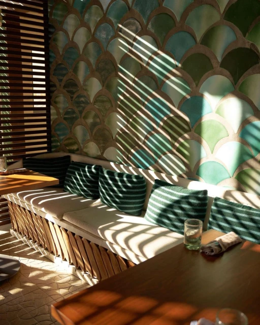 Indoor seating area with turquoise and green patterned wall, wooden benches, and pillows, illuminated by natural light streaming through blinds.