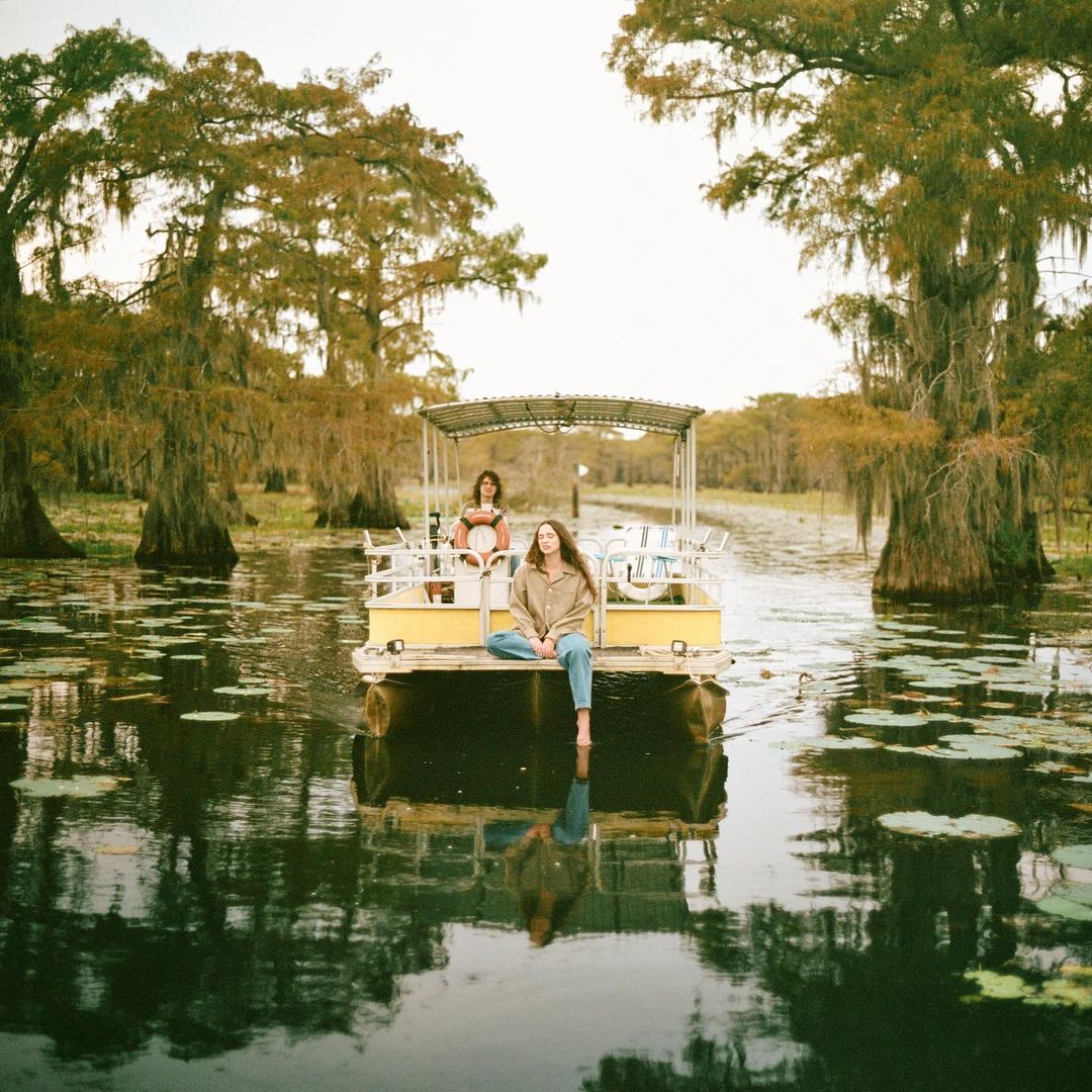 Two adult women riding on a yellow pontoon boat along a river in a swampy, wooded area on a hazy day with trees overhead.