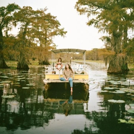 Two adult women riding on a yellow pontoon boat along a river in a swampy, wooded area on a hazy day with trees overhead.