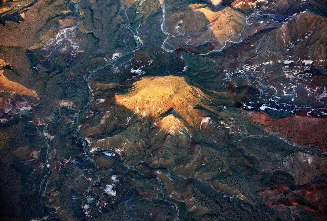 Aerial view of a mountain range with rivers and valleys, bathed in golden sunlight, creating a textured landscape with deep shadows.