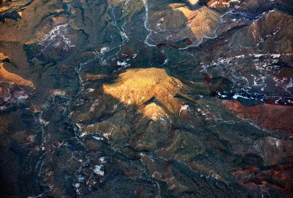 Aerial view of a mountain range with rivers and valleys, bathed in golden sunlight, creating a textured landscape with deep shadows.