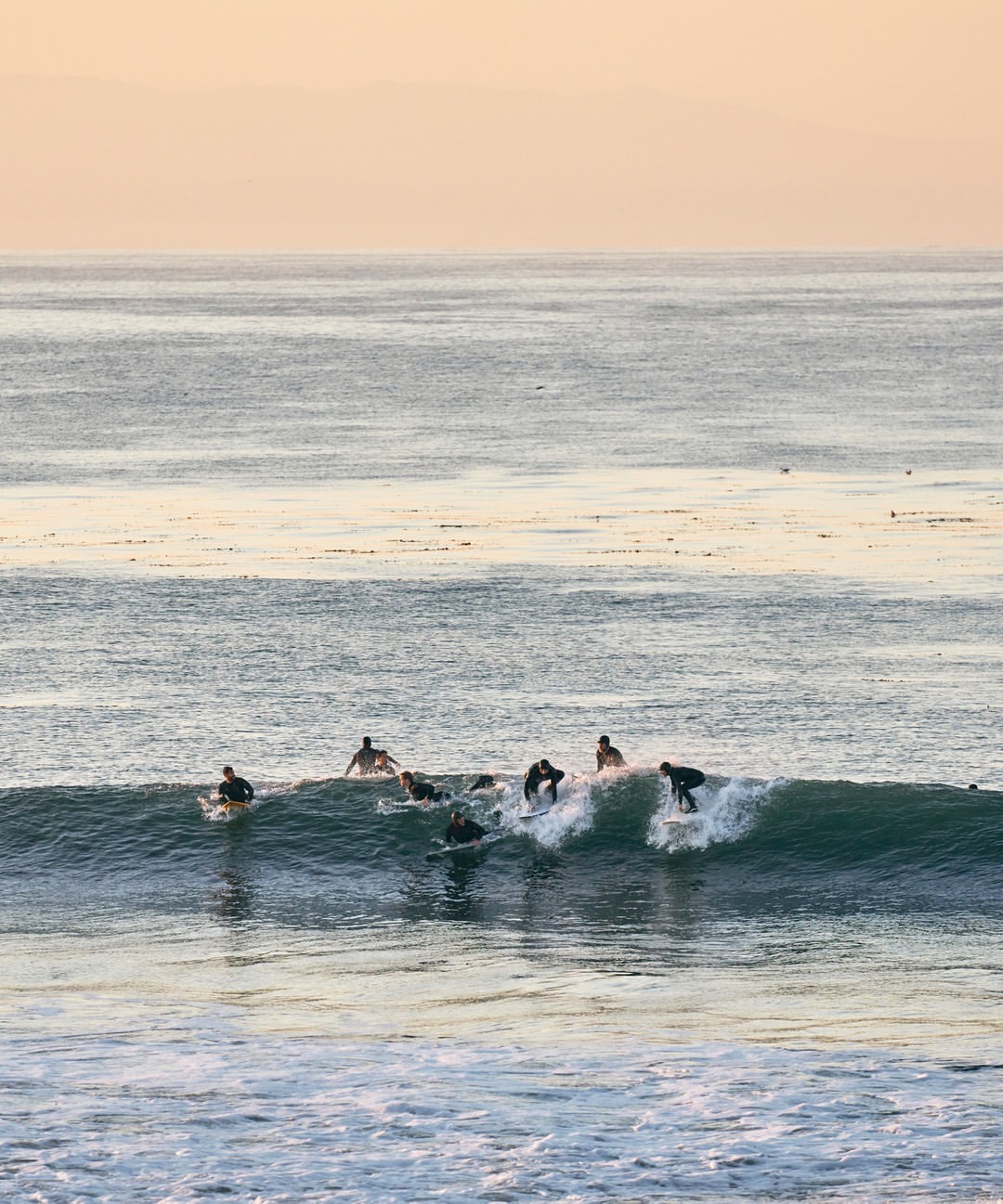 Several surfers ride a wave in the ocean on their surfboards, silhouetted against the sunset, creating an athletic watersport scene.