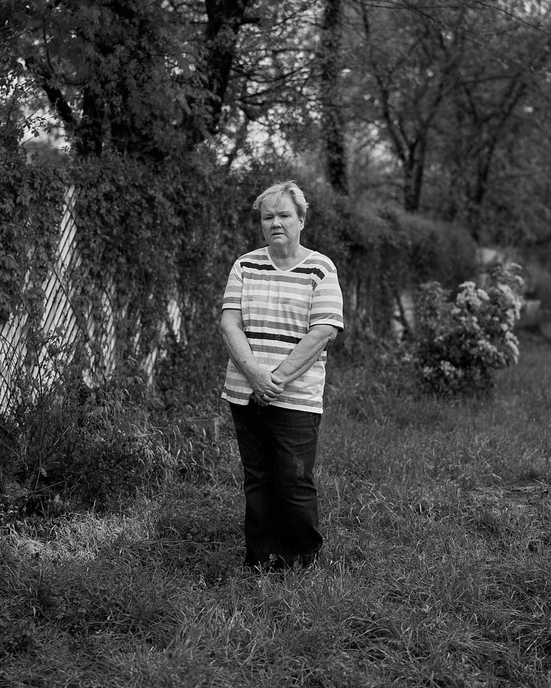 Black-and-white portrait of a mature adult woman standing in a grassy field, wearing a striped shirt, with a foliage-covered fence backdrop.