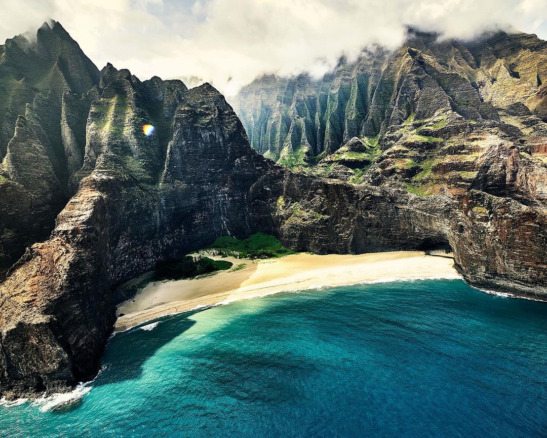Aerial view of rugged mountains meeting the ocean at a sandy beach, with the blue water contrasting against the dark rocks and cliffs.