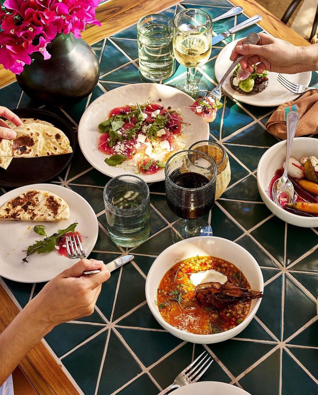 Overhead shot of a dinner table setting with multiple white plates filled with food and several glasses of drinks, and pink flowers.