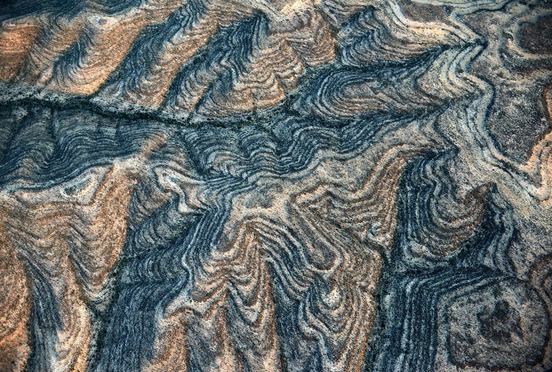 Abstract full-frame view of banded gneiss rock formations, featuring wavy lines and varying shades of grey, blue, and tan colors.