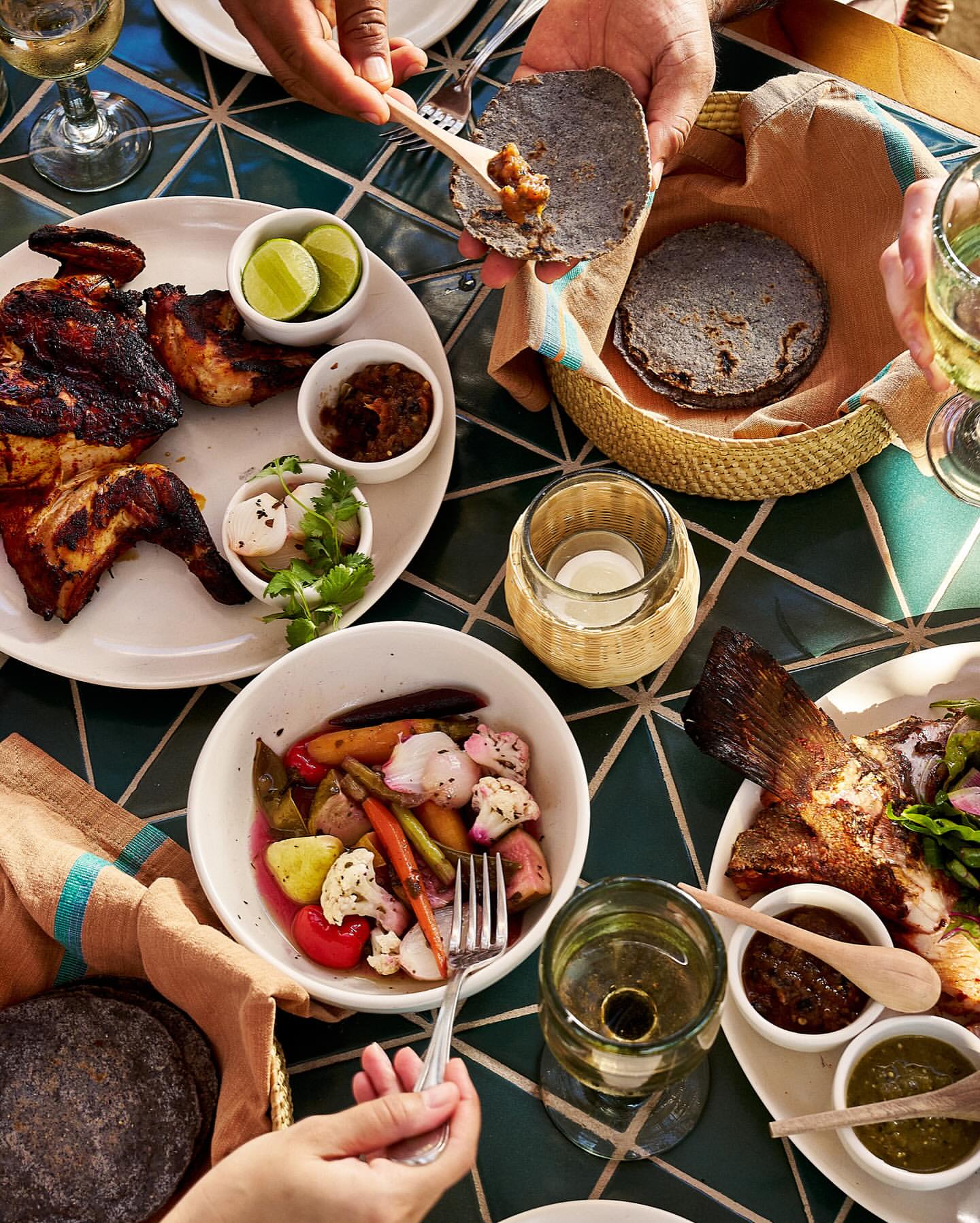 Overhead shot of a table with multiple plates of food including grilled chicken, fish, vegetables, tortillas and drinks, hands reaching for food.