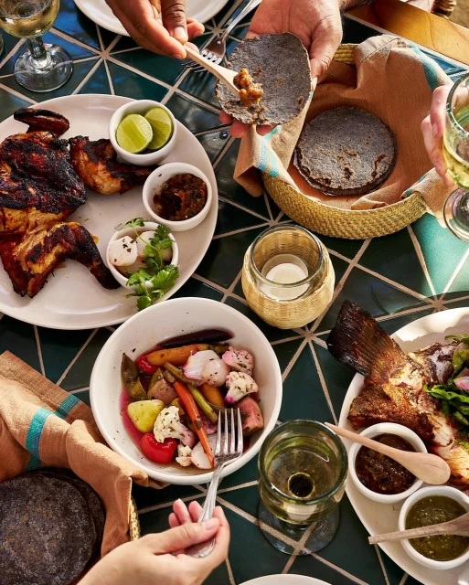 Overhead shot of a table with multiple plates of food including grilled chicken, fish, vegetables, tortillas and drinks, hands reaching for food.