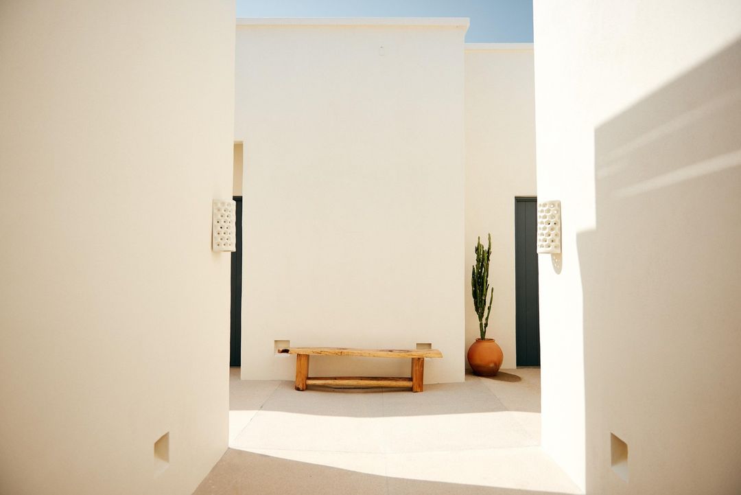 Architectural scene features a light wooden bench, potted cactus, and symmetrical walls with shadow patterns in an open-air courtyard.