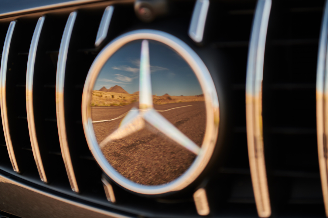 Close-up of a luxury car grill reflecting a desert road and mountains in the car's logo emblem, captured in warm, natural lighting.