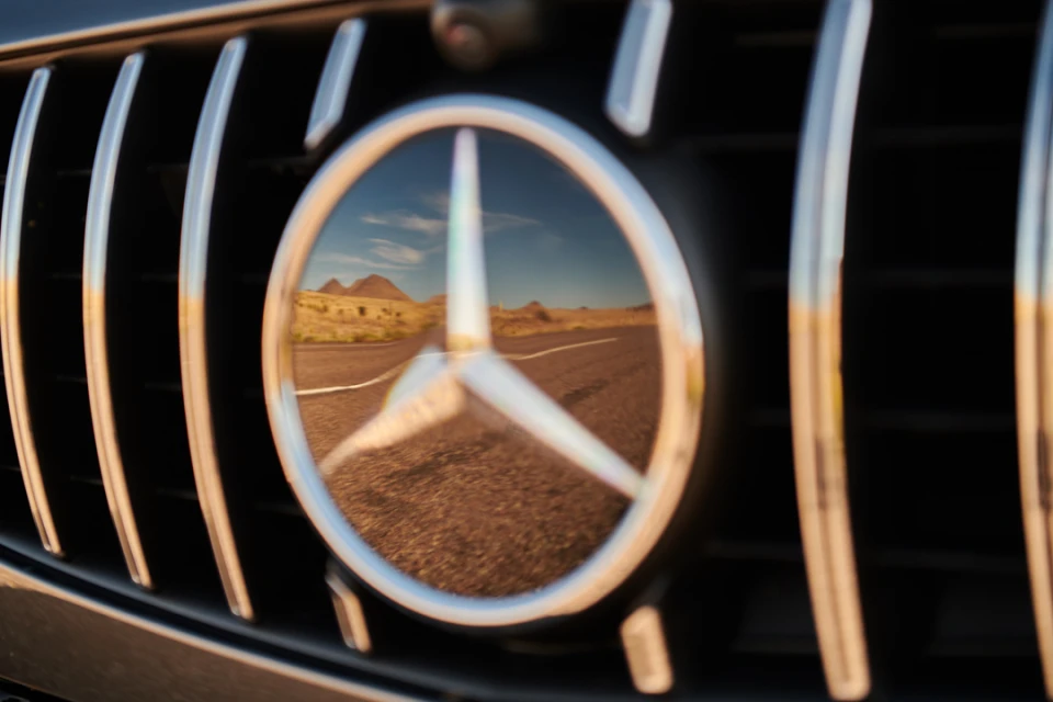 Close-up of a luxury car grill reflecting a desert road and mountains in the car's logo emblem, captured in warm, natural lighting.