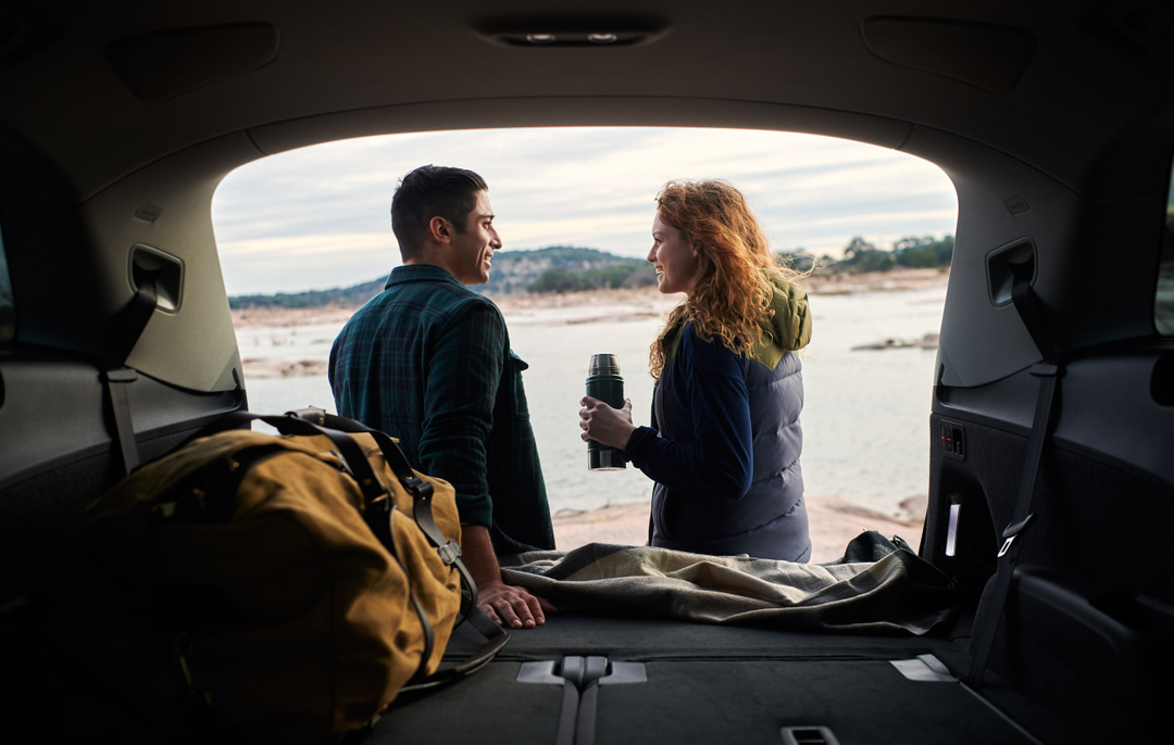 A couple in the back of an SUV looks out over a lake, with a thermos, blanket, and duffel bag visible inside the vehicle.