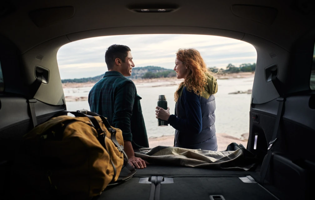 A couple in the back of an SUV looks out over a lake, with a thermos, blanket, and duffel bag visible inside the vehicle.