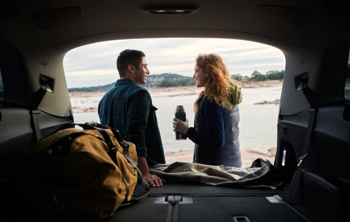 A couple in the back of an SUV looks out over a lake, with a thermos, blanket, and duffel bag visible inside the vehicle.
