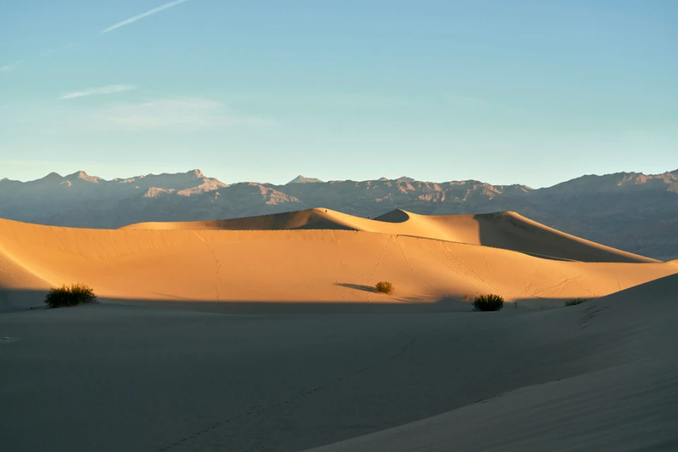 Desert landscape with sand dunes and distant mountains under a blue sky. Golden light illuminates the dunes in the foreground.