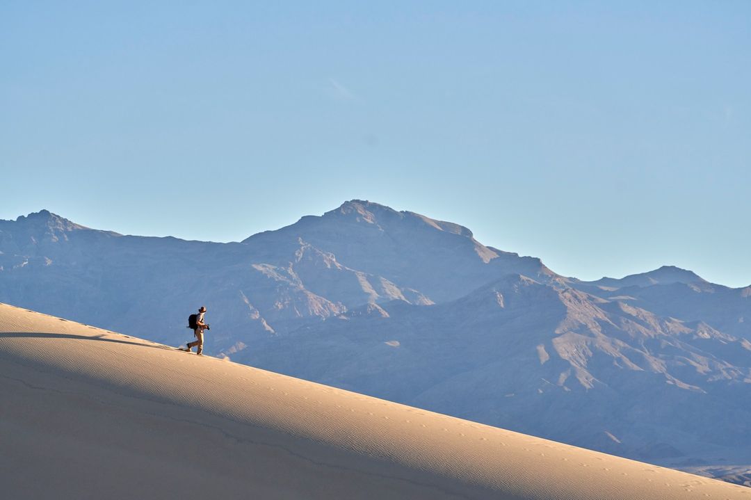 Adult hiker treks up a large sand dune with distant mountains under a clear blue sky in a wide outdoor shot, capturing a nature scene.