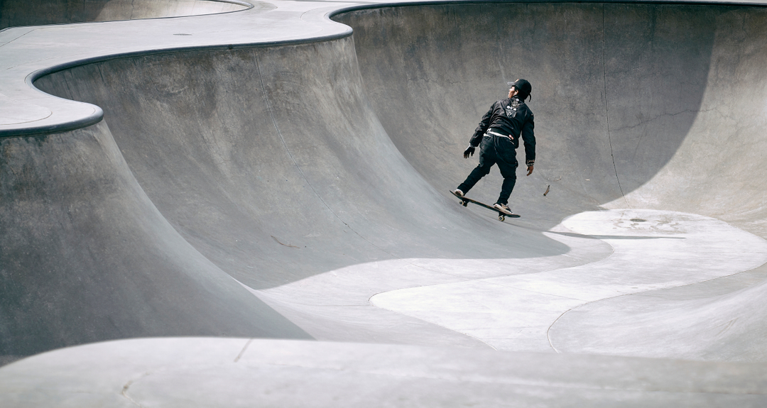 An adult male in dark athletic apparel skateboarding on a curved concrete surface in a skatepark on a sunny day.
