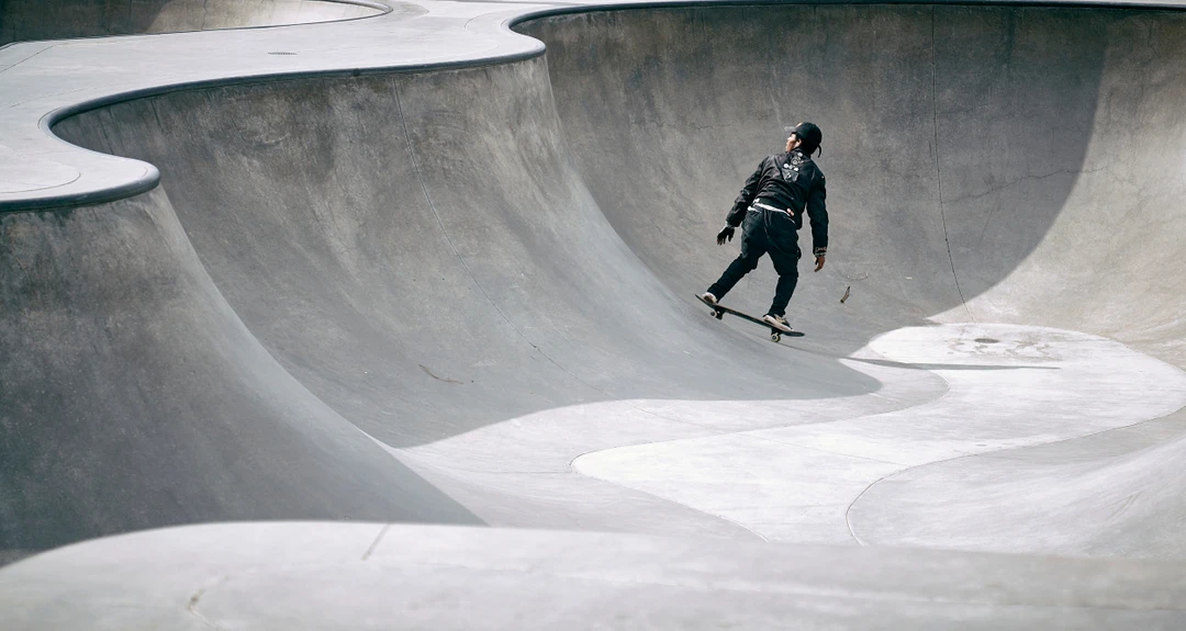 An adult male in dark athletic apparel skateboarding on a curved concrete surface in a skatepark on a sunny day.