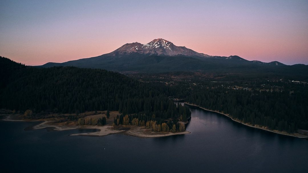 Aerial view of Mount Shasta with snow-capped peak and forests surrounding a blue lake, during the pastel colors of sunset.