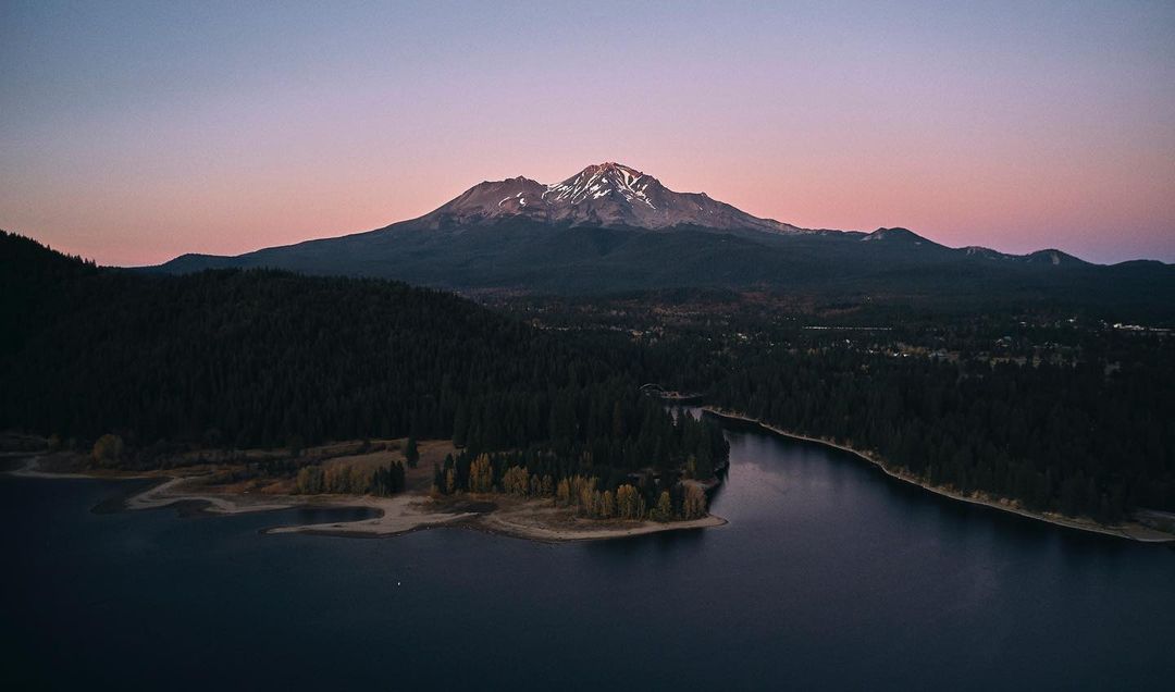 Aerial landscape view of Mount Shasta reflecting on a lake surrounded by a dark green forest during a blue and pink hued dusk.