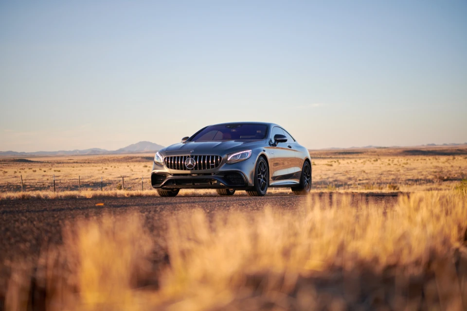 Gray Mercedes-Benz coupe parked in a grassy desert landscape, captured in a low-angle shot during the daytime with a clear sky.