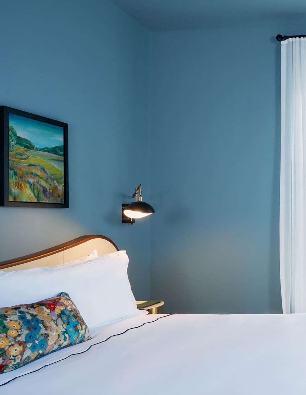 Bedroom interior featuring a bed with white linens and floral pillow, a painting, a lamp, and a window with sheer white curtains.