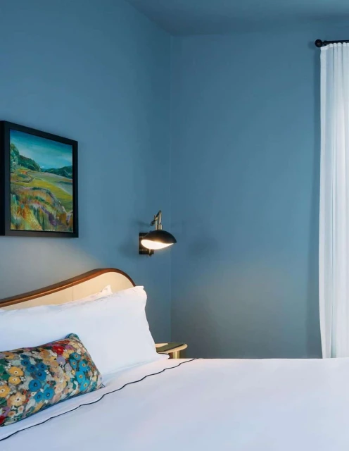Bedroom interior featuring a bed with white linens and floral pillow, a painting, a lamp, and a window with sheer white curtains.