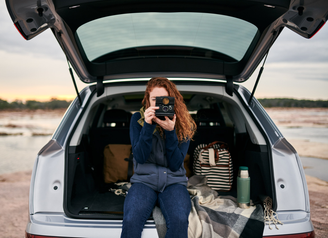 A woman with red hair sits in the back of a silver SUV taking a photo with a vintage Polaroid camera in an outdoor setting.