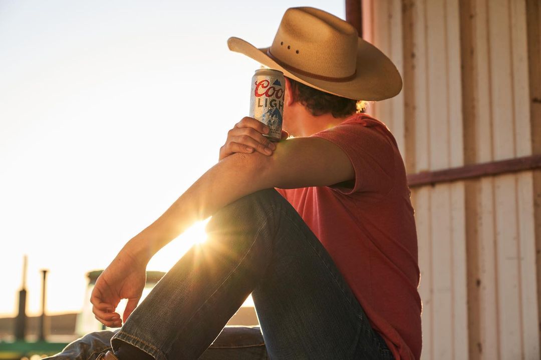 Man in cowboy hat sits in the sun with a can of Coors Light beer in hand wearing jeans and a red shirt, resting on his elbow.