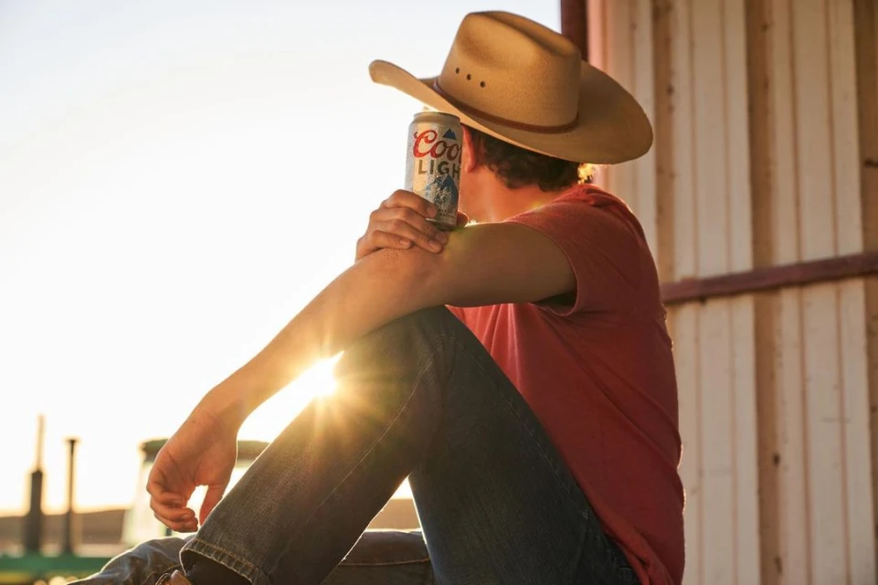 Man in cowboy hat sits in the sun with a can of Coors Light beer in hand wearing jeans and a red shirt, resting on his elbow.