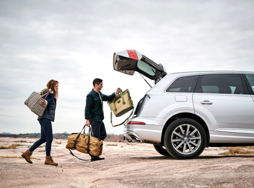 Couple loading luggage into a silver Audi SUV outdoors against a cloudy sky on a rocky surface, preparing for travel or road trip.
