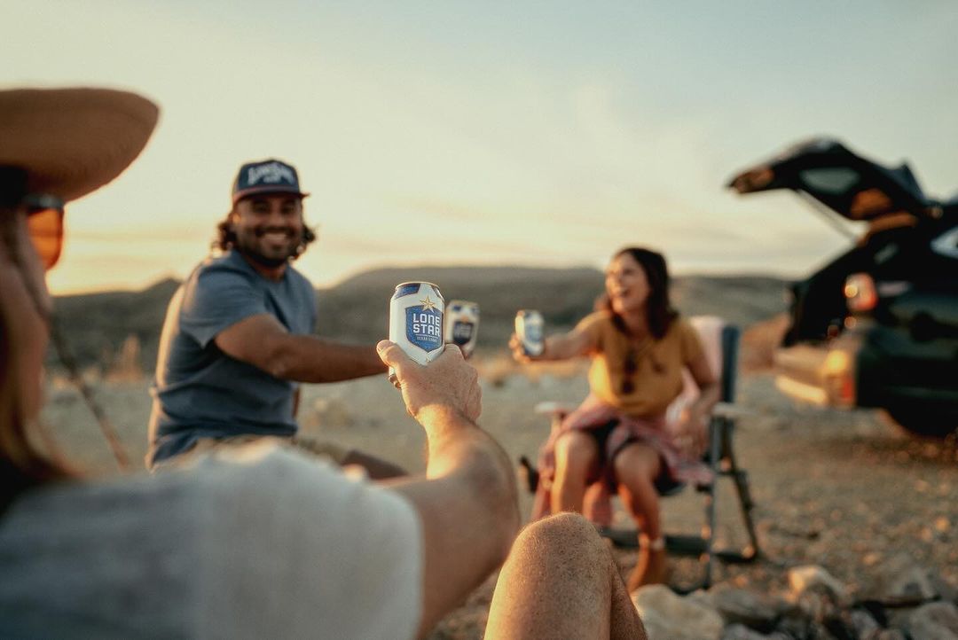 Friends toasting with Lone Star beer cans at sunset, outdoor gathering, casual wear, car in background, relaxed desert atmosphere.