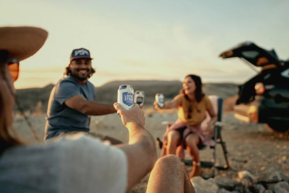 Friends toasting with Lone Star beer cans at sunset, outdoor gathering, casual wear, car in background, relaxed desert atmosphere.