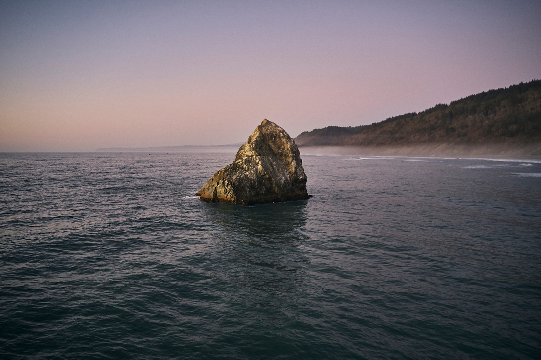 Ocean landscape featuring a prominent rock formation in the water, with a forested coastline and a dusky sky at dawn or dusk.