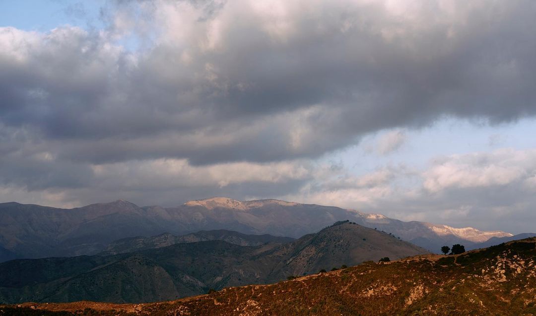 Hills and mountains in a vast landscape are partially lit with the sky filled with heavy gray clouds overhead in an outdoor scene.