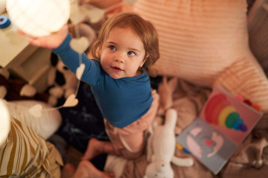 Toddler in blue shirt and pink pants kneels surrounded by pillows, toys, and book in a cozy indoor setting.