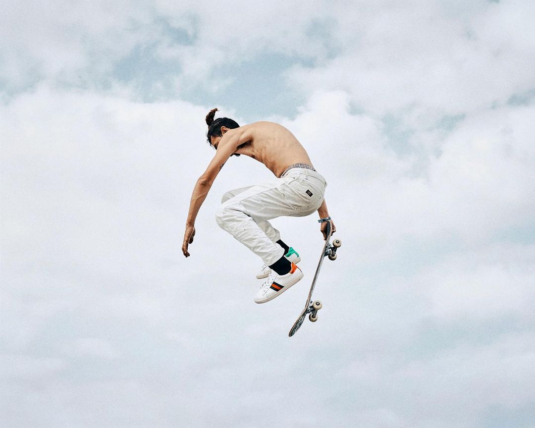 A shirtless man with a topknot performs a mid-air skateboard trick against a cloudy sky, wearing white pants and multicolored sneakers.