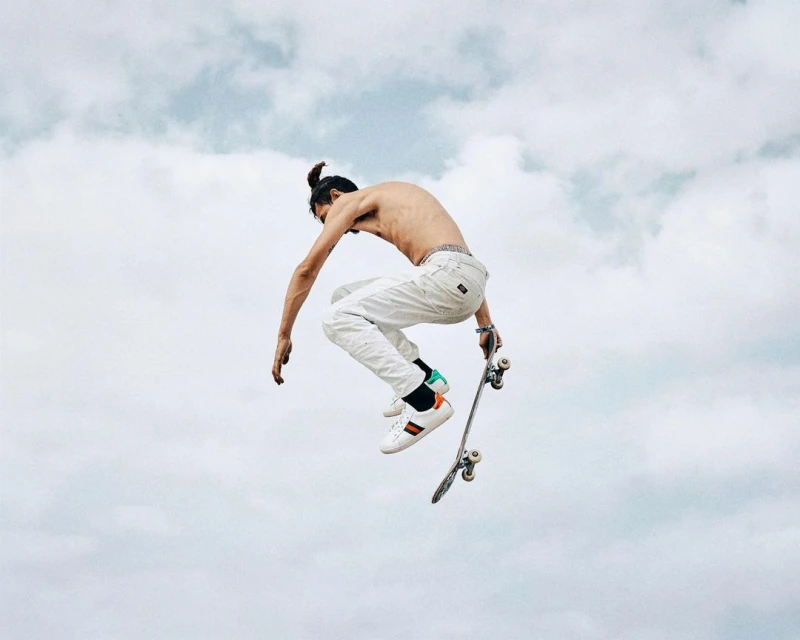 A shirtless man with a topknot performs a mid-air skateboard trick against a cloudy sky, wearing white pants and multicolored sneakers.