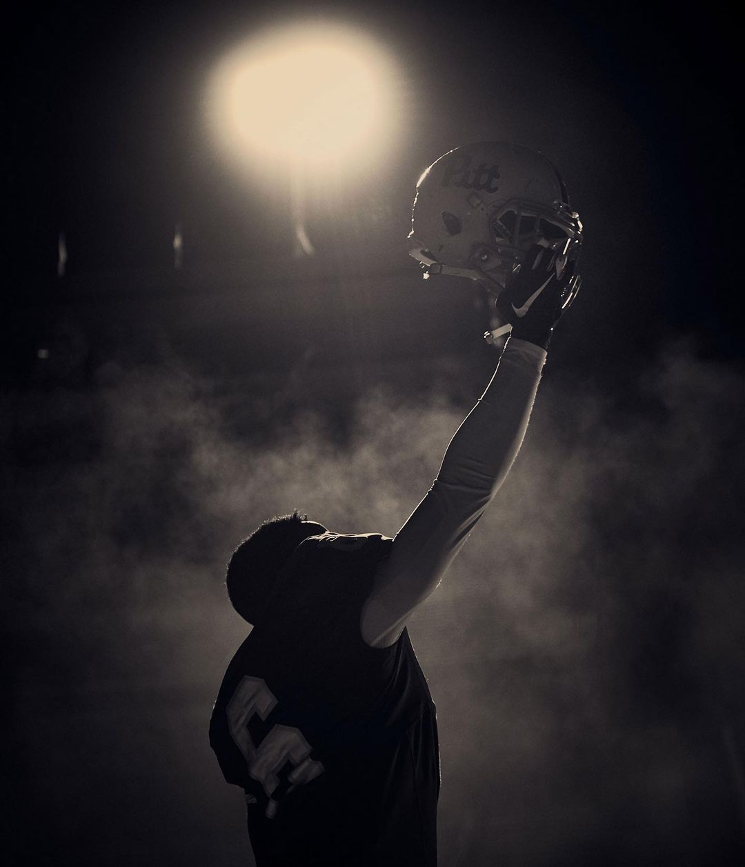 A person in a dark environment holding a football helmet up toward a bright light in a sepia-toned image.
