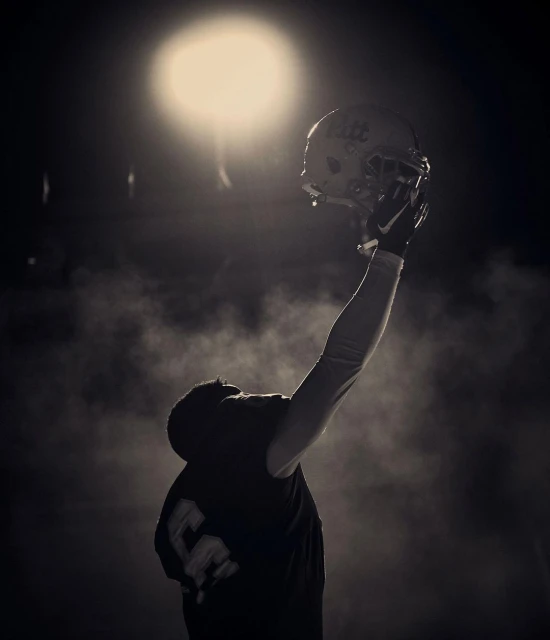 A person in a dark environment holding a football helmet up toward a bright light in a sepia-toned image.