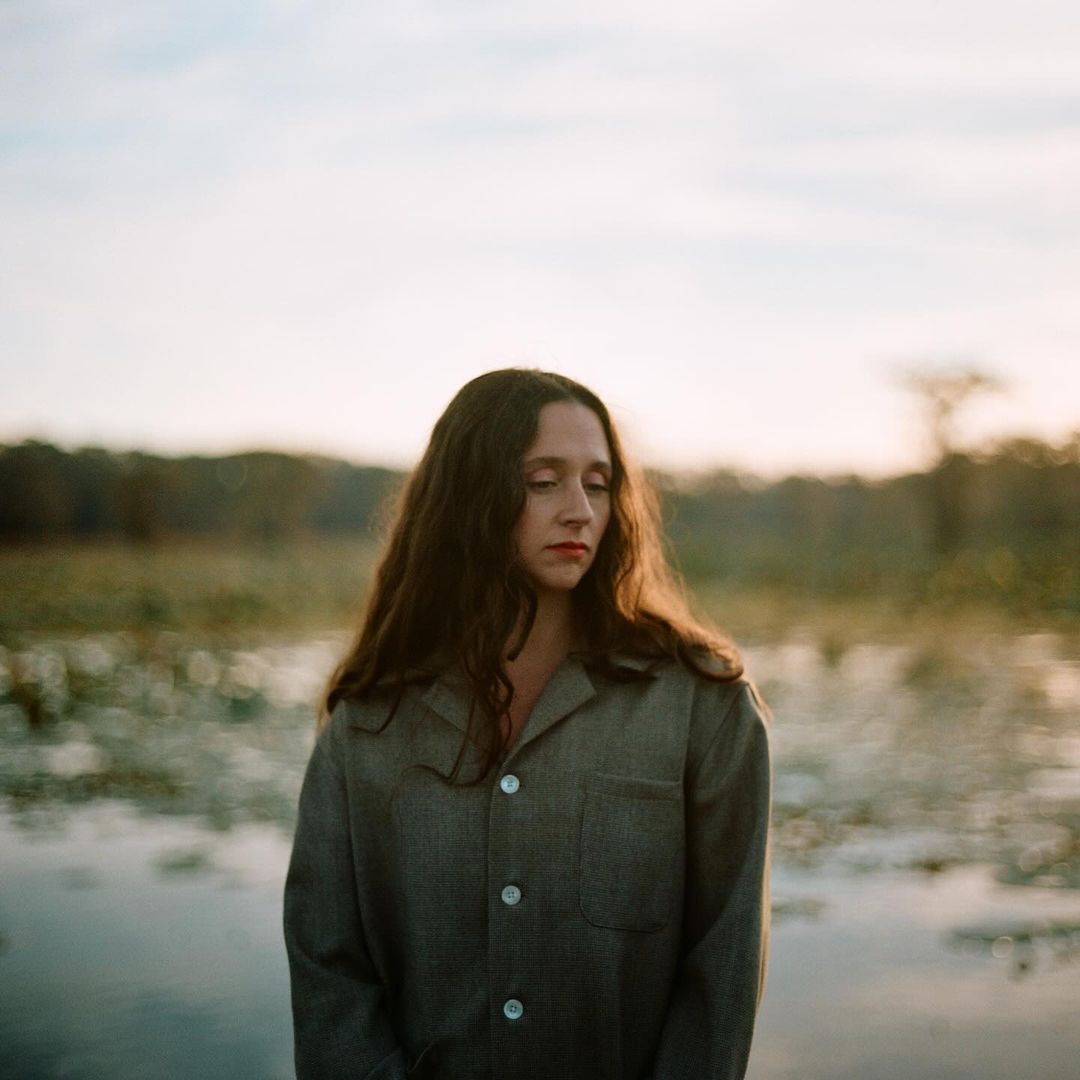 Portrait of a contemplative woman with long dark hair, wearing a grey button-down shirt, standing outdoors near a body of water.