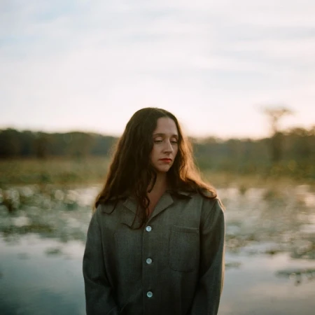 Portrait of a contemplative woman with long dark hair, wearing a grey button-down shirt, standing outdoors near a body of water.